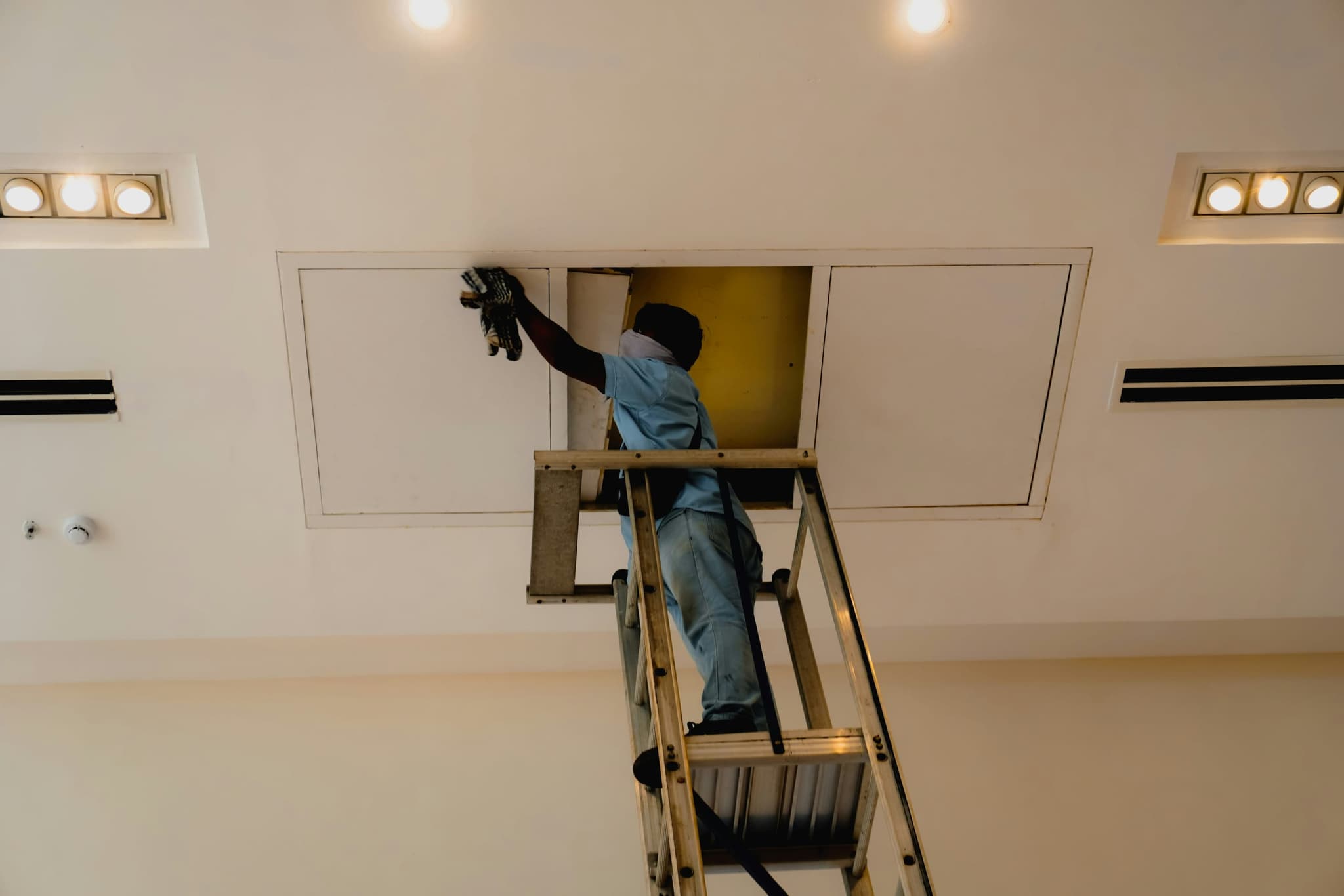 A technician on a ladder opening a ceiling hatch to reach the equipment above it
